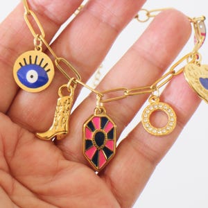 Könnte beinhalten: Ein goldfarbenes Gliederkettenarmband mit mehreren hängenden Anhängern, darunter ein blau-weißes Auge, ein Cowboystiefel, ein geometrischer Anhänger in Pink und Schwarz, ein runder Strassanhänger und ein herzförmiger Anhänger mit blauem Kern.