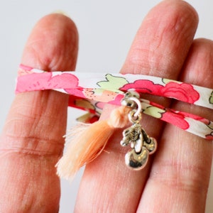 May include: A double-layered fabric bracelet with a floral print in pink and white. The bracelet has a silver charm with a peach-coloured tassel and a silver cherry charm.