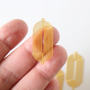 Peut inclure: Composants de boucles d'oreilles géométriques dorés. Les composants ont une forme ovale allongée avec une série de lignes courbes à l'intérieur. Deux petits trous sont en haut et en bas pour la fixation. Les composants sont tenus dans une main.