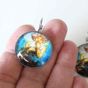 Peut inclure: Boucles d'oreilles rondes argentées avec un motif Art nouveau vintage. Chaque boucle d'oreille présente une image détaillée d'une femme aux cheveux flottants, sur fond bleu et or. Fermeture à levier.
