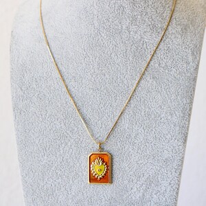 May include: A gold-toned necklace featuring a rectangular pendant with a burnt orange background and a central yellow heart emblem. The heart is surrounded by a decorative scalloped border and a small clear stone at its center, suspended on a thin chain.