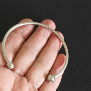 Könnte beinhalten: Ein silbernes Armreif-Armband mit zwei runden Kugelenden.