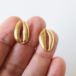 Peut inclure: Deux boucles d'oreilles dorées en forme de coquillage. Les boucles d'oreilles sont ovales avec un intérieur détaillé et strié. Les boucles d'oreilles sont tenues dans la main d'une personne sur fond blanc.