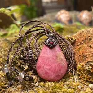 May include: A pink teardrop-shaped pendant necklace with a brown woven cord. The pendant is a vibrant pink with a textured surface. The necklace is displayed on a mossy surface with a blurred natural background. The pendant is approximately 1.5 inches in length.
