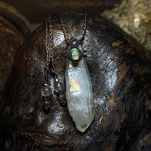 May include: A handmade necklace with a clear quartz crystal pendant. The pendant hangs from a brown cord, with an iridescent opal detail. The necklace has a natural, earthy look, highlighting the crystal's clarity and unique form. The pendant is approximately 10 cm long.