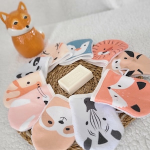 Może przedstawiać: Kolekcja myjek z motywami zwierzęcymi ułożonych wokół kostki mydła na plecionej macie. Myjki przedstawiają różne zwierzęce twarze w miękkich kolorach. Ceramiczna figurka lisa znajduje się w tle.