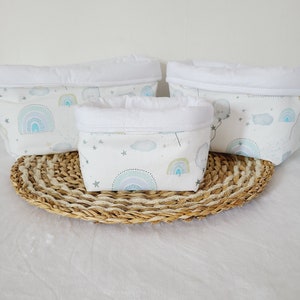 May include: Three white fabric storage baskets with a white trim. The baskets have a light blue and green pattern with rainbows, clouds, and stars. The baskets are sitting on a woven straw mat.