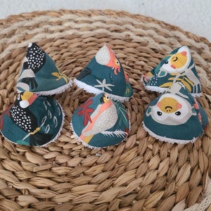 May include: Six teal fabric items with bird and floral patterns, each with a white trim. The items are arranged on a woven, circular surface. The bird designs feature various colors, including orange, yellow, and white.