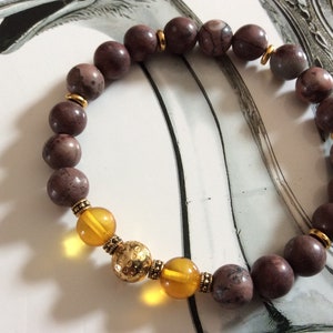 May include: A brown and gold beaded bracelet with a textured gold bead in the center. The bracelet is made with natural stones and has a simple design.