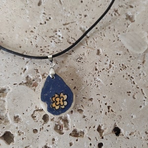 Peut inclure: Un collier pendentif en forme de goutte d'eau avec un motif bleu foncé et crème. Le pendentif présente un motif de fleur jaune. Le collier a un cordon noir et un fermoir argenté.