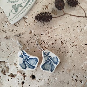 Peut inclure: Boucles d'oreilles artisanales avec un motif floral bleu sur des pièces de céramique blanche. Les boucles d'oreilles sont suspendues à des crochets argentés. Le fond est une surface texturée et claire avec des éléments décoratifs.