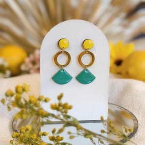 Può includere: Orecchini con un cerchio giallo, un anello color oro e una forma a ventaglio turchese. Gli orecchini sono esposti su un supporto bianco, con uno specchio e fiori gialli sullo sfondo.
