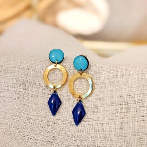 Può includere: Un paio di orecchini pendenti con una parte superiore circolare blu turchese, una parte centrale circolare color oro e una parte inferiore a forma di diamante blu navy. Gli orecchini sono esposti su una superficie bianca strutturata.
