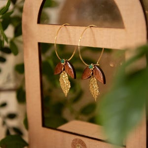 May include: Gold hoop earrings with dark wood and gold leaf details. Each earring has a small green gem. The earrings are displayed on a wooden stand with the text "by Héloïse".