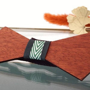 Op de afbeelding: Houten vlinderdas in een diepe roodachtig bruine tint. Het heeft een zwarte band met een groen en wit chevron patroon. Een stijlvol accessoire voor een formele of semi-formele gelegenheid.
