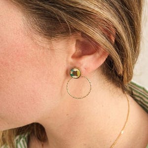 May include: Gold-coloured hoop earrings with a textured finish. Each earring has a small, circular stud with a colourful, abstract design. A delicate gold chain necklace is also visible.