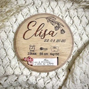 Puede incluir: Una placa redonda de madera con un diseño floral y el nombre "Elisa" grabado en ella. La placa también incluye la fecha "22.01.2020", la hora "23h44", la longitud "55 cm" y el peso "4kg150".