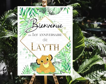 May include: A welcome sign for a first birthday party with a jungle theme. The sign features a green and gold diamond shape with the text "Bienvenue au 1er Anniversaire de LAYTH" and a cartoon lion cub.