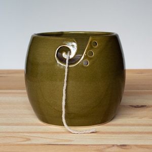 Puede incluir: Cuenco de cerámica verde oliva para hilo con una guía en espiral y tres pequeños agujeros. Un hilo de lana blanquecina sale del cuenco. El cuenco está sobre una superficie de madera clara.