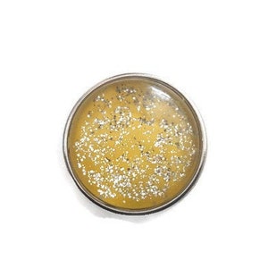 Puede incluir: Un charm redondo plateado con fondo amarillo y purpurina plateada.