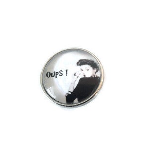 May include: A round, silver-colored button with a black and white image of a woman with the text "Oups!" printed on it.