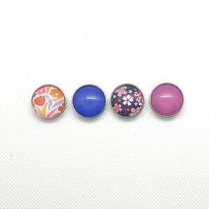 Puede incluir: Cuatro botones redondos a presión con diferentes diseños. Un botón tiene un patrón floral rosa, uno tiene un color sólido azul, uno tiene un color sólido rosa y uno tiene un patrón floral multicolor.