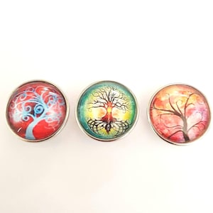 Könnte beinhalten: Drei runde, silberfarbene, dekorative Knöpfe mit bunten Baummotiven. Der erste Knopf hat einen roten Hintergrund mit einem blauen und weißen Baum. Der zweite Knopf hat einen grünen Hintergrund mit einem braunen Baum. Der dritte Knopf hat einen rosa Hintergrund mit einem braunen Baum.