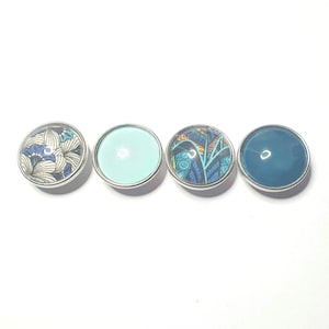 Puede incluir: Cuatro botones a presión redondos con diferentes diseños. El primer botón tiene un fondo blanco con un patrón floral azul y blanco. El segundo botón es azul claro sólido. El tercer botón tiene un diseño estampado azul y blanco. El cuarto botón es azul oscuro sólido.