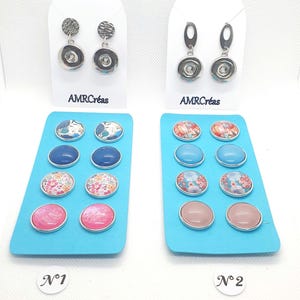 Puede incluir: Se muestran dos pares de pendientes y dos juegos de botones decorativos. Los pendientes tienen un disco plateado texturizado y un círculo plateado más pequeño. Los botones presentan patrones florales y colores lisos. El texto "AMRCréas" es visible.