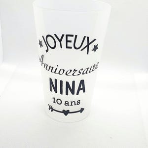 Może przedstawiać: Biały plastikowy kubek z czarnym tekstem, który brzmi "Joyeux Anniversaire Nina 10 ans" z czarnym wzorem strzałki i serca.