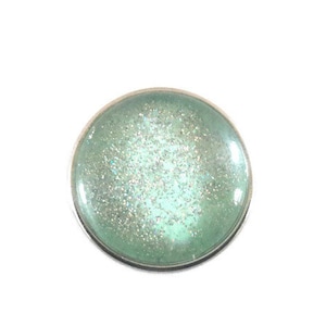 Puede incluir: Un charm redondo con borde plateado y relleno de resina azul claro brillante.