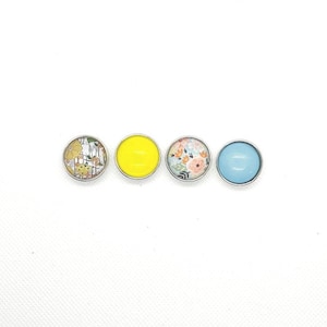 Puede incluir: Cuatro botones redondos con borde plateado y diseños diferentes. Un botón es amarillo sólido, uno tiene un patrón floral, uno tiene un patrón a rayas azul y blanco, y uno es azul sólido.