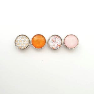 May include: Four round, silver-rimmed buttons with different designs. One button has a white and gold geometric pattern, one has a solid orange color, one has a white and pink floral pattern, and one is solid pink.