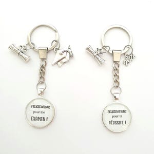 Peut inclure: Deux porte-clés argentés avec des breloques sur le thème de la graduation. Un porte-clés a un charme avec le texte "Felicitations pour ton examen!" et l'autre porte-clés a un charme avec le texte "Felicitations pour ta reussite!"