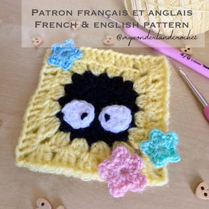 Może przedstawiać: Kwadratowy szydełkowy w kolorze żółtym z czarną postacią i białymi oczami pośrodku. Małe szydełkowe elementy w kształcie gwiazd w kolorze niebieskim i różowym są umieszczone wokół kwadratu. Widoczny jest tekst "FRENCH & ENGLISH PATTERN".