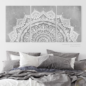 May include: Three-panel wall art featuring a white mandala design on a gray background. The intricate pattern is displayed above a bed with gray and white bedding. The artwork adds a touch of elegance to the bedroom decor.