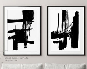 May include: Two black and white abstract art prints with thick brushstrokes, hanging on a light gray wall above a gray couch with gray throw pillows and a gray throw blanket. The couch is in a living room with a wooden floor, two wooden stools, a small round table, and a black floor lamp with two shades.