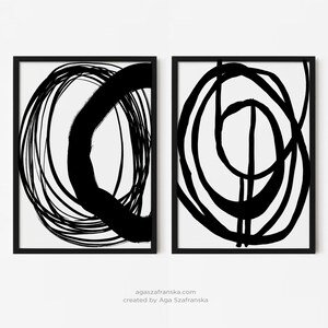 Op de afbeelding: Twee ingelijste abstracte kunstprints. Elke print heeft dikke zwarte penseelstreken op een witte achtergrond. Het kunstwerk bevat cirkelvormige en lineaire vormen. De frames zijn zwart. De website en de naam van de kunstenaar staan onderaan.