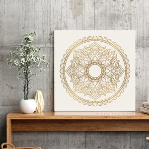 May include: A square canvas print with a white background and a gold mandala design. The mandala is intricate and detailed, with a circular pattern of overlapping petals and leaves. The canvas is framed with a thin white border.