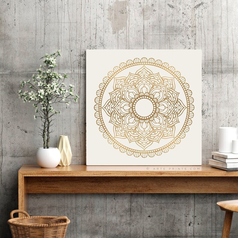 May include: A square canvas print with a white background and a gold mandala design. The mandala is intricate and detailed, with a circular pattern of overlapping petals and leaves. The canvas is framed with a thin white border.