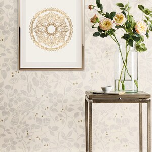 May include: A silver metal end table with a glass vase filled with pink roses and a small white bowl on top. The table is in front of a white wall with a floral pattern and a framed gold mandala print.