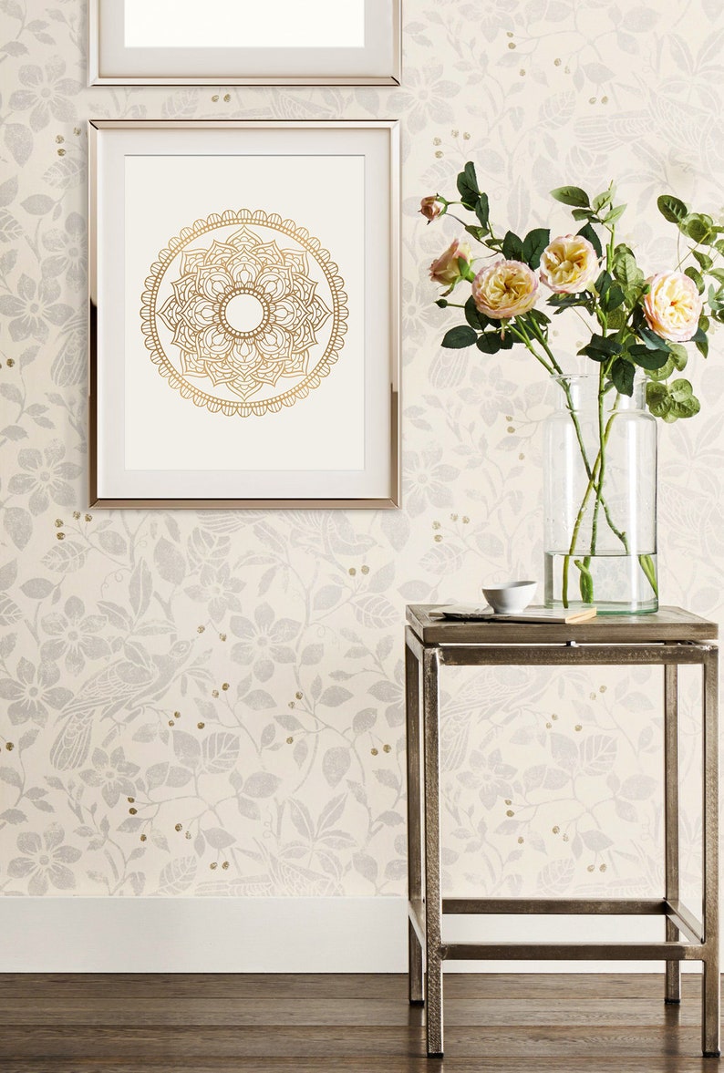 May include: A silver metal end table with a glass vase filled with pink roses and a small white bowl on top. The table is in front of a white wall with a floral pattern and a framed gold mandala print.