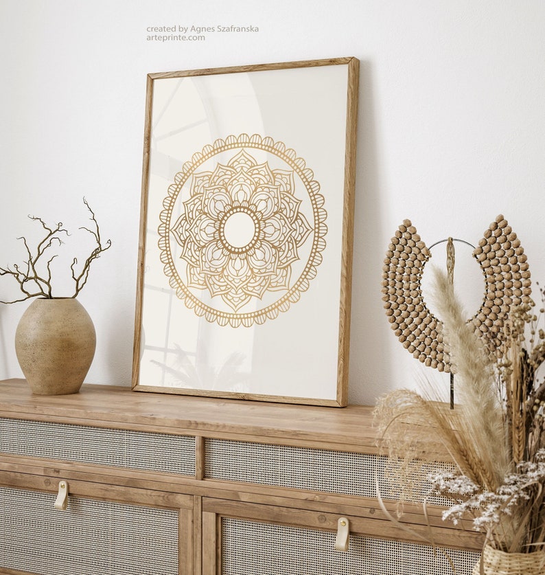 May include: A framed print of a gold mandala design on a white background. The print is displayed on a wooden shelf with a vase of dried branches and a wooden beaded necklace.