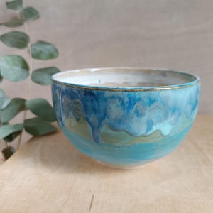 Op de afbeelding: Handgemaakte keramische kom met een glanzend, druipend glazuur in blauwe en turquoise tinten. De kom heeft een lichtbeige binnenkant en een gouden rand. De kom staat op een houten oppervlak. Diameter ongeveer 15 cm.
