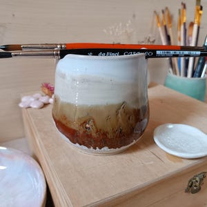Peut inclure: Une tasse en céramique faite à la main avec un glaçage blanc, beige et marron, contenant deux pinceaux. Les pinceaux ont des poils noirs et des manches orange et noirs. Une petite coupelle blanche est à proximité, avec d'autres fournitures d'art.