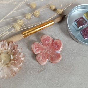 Peut inclure: Gros plan sur une fleur en céramique émaillée rose et blanche. Un pinceau à manche doré et une petite boîte métallique contenant des aquarelles sont également visibles. Des fleurs séchées sont en arrière-plan.