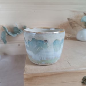 Può includere: Una tazza in ceramica fatta a mano con una forma arrotondata. La tazza presenta una glassa con sfumature di blu, verde e bianco, creando un effetto acquerello. Il bordo è di colore marrone chiaro. La tazza è appoggiata su una superficie di legno.