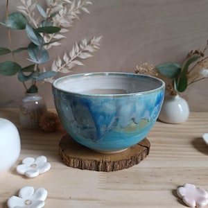 Könnte beinhalten: Handgefertigte Keramikschale mit blau-türkisfarbener Glasur. Die Schale steht auf einer Holzscheibe. Innen weiß, außen hellblau. Dekorative Blumen und Vasen im Hintergrund.