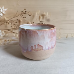 Könnte beinhalten: Ein Keramikbecher mit abgerundeter Form, der eine glatte, hellbeige Basis aufweist, die in eine glänzende, tropfende Glasur in zarten Rosa- und Weißtönen übergeht. Das Innere des Bechers ist mit einer glatten, hellen Glasur versehen.