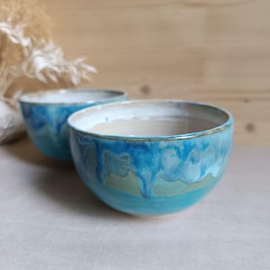 Peut inclure: Deux bols en céramique présentant une glaçure brillante avec un motif dégoulinant dans des tons turquoise, bleu profond et vert clair. L'intérieur de chaque bol est d'une couleur blanc cassé neutre et lisse, avec des bords finis par un liseré métallique.